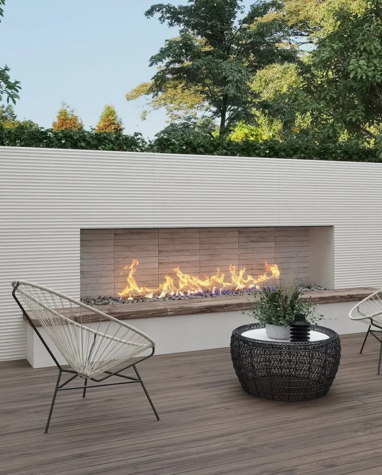 Modern organic patio with a long built-in fireplace, two wire-frame chairs, a round wicker table, and trees in the background.