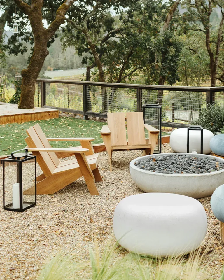 Two wooden chairs face a round stone fire pit, surrounded by white seating poufs, lanterns, and trees in a fenced outdoor patio area with an inviting organic modern vibe.