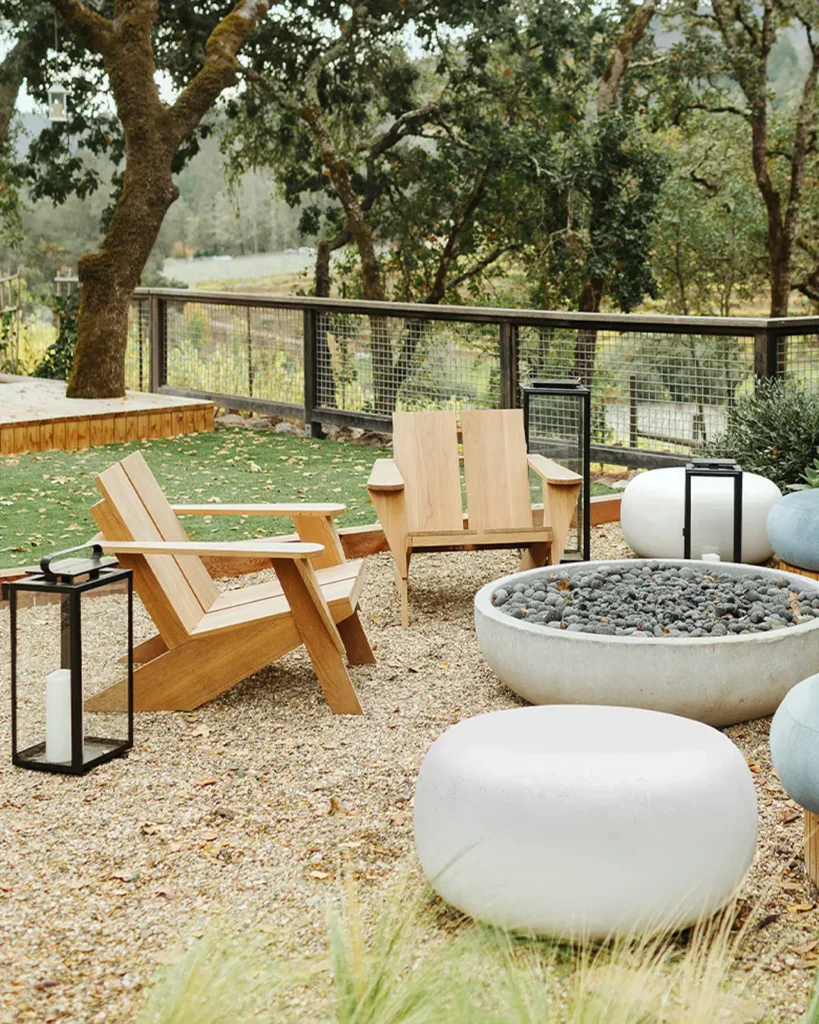Two wooden chairs face a round stone fire pit, surrounded by white seating poufs, lanterns, and trees in a fenced outdoor patio area with an inviting organic modern vibe.