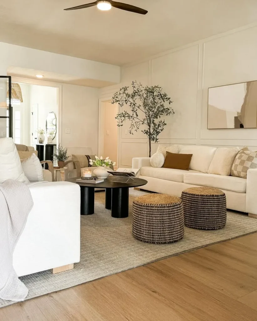 A modern living room blends organic modern vs minimalist styles, featuring neutral-toned furniture, round black coffee tables, woven ottomans, a light rug, wood floor, and a potted tree near a beige sofa.