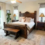 Rustic bedroom with a wooden bed featuring a carved headboard and matching bench, brightened by two nightstands, lamps, a potted plant, rug, and large windows with light curtains.