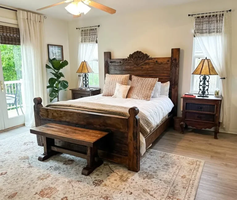 Rustic bedroom with a wooden bed featuring a carved headboard and matching bench, brightened by two nightstands, lamps, a potted plant, rug, and large windows with light curtains.