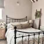 A neatly made bed with a floral quilt and beige pillows sits amid rustic bedroom furniture in a cozy space with exposed wooden beams, a wooden ladder draped with a blanket, and a small bedside table topped with flowers.