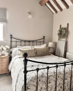 A neatly made bed with a floral quilt and beige pillows sits amid rustic bedroom furniture in a cozy space with exposed wooden beams, a wooden ladder draped with a blanket, and a small bedside table topped with flowers.
