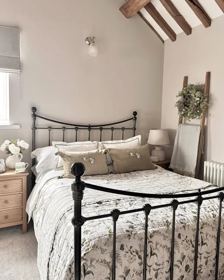 A neatly made bed with a floral quilt and beige pillows sits amid rustic bedroom furniture in a cozy space with exposed wooden beams, a wooden ladder draped with a blanket, and a small bedside table topped with flowers.