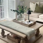 A rustic coffee table decor features a wooden table with a green throw, woven trays, a glass vase with greenery, a candle, and a lamp, set in a neutral-toned living room with a sofa and large window shutters.