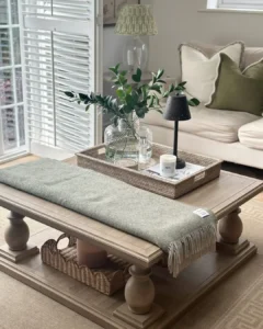 A rustic coffee table decor features a wooden table with a green throw, woven trays, a glass vase with greenery, a candle, and a lamp, set in a neutral-toned living room with a sofa and large window shutters.