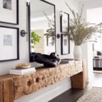 A rustic console table with decorative objects and a large vase of branches stands against a wall adorned with framed art, a rectangular mirror, and two wall sconces.