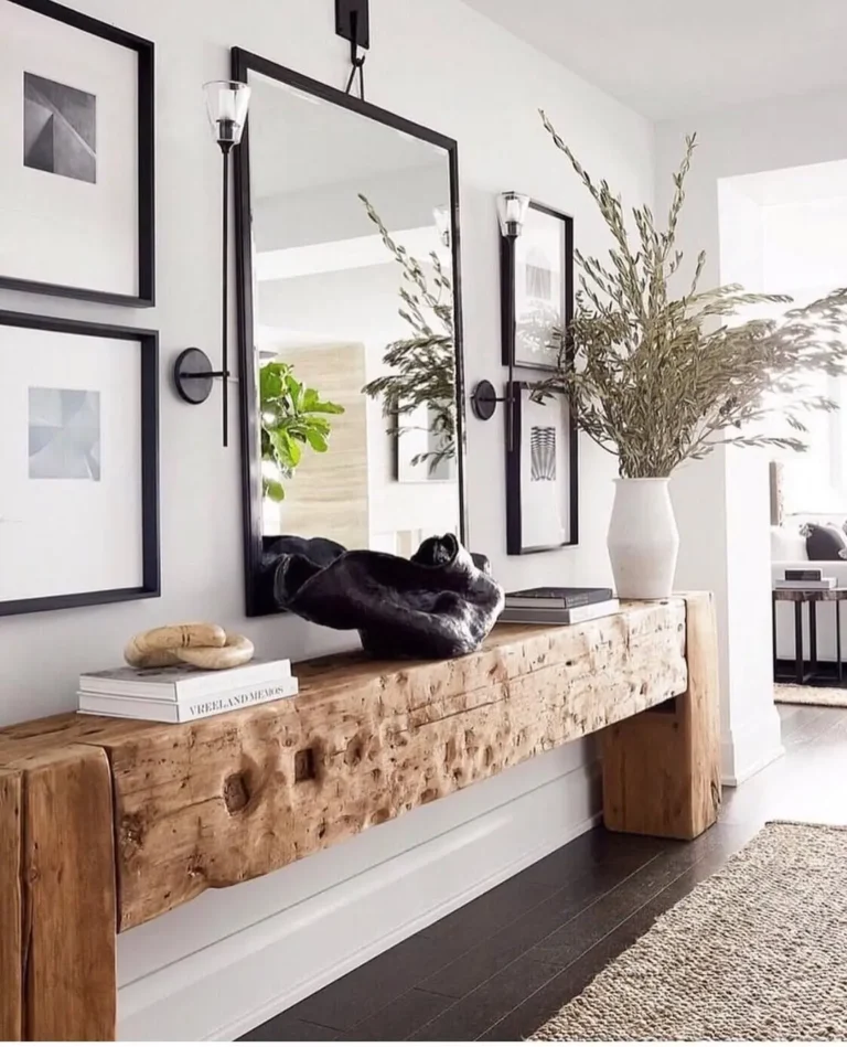 A rustic console table with decorative objects and a large vase of branches stands against a wall adorned with framed art, a rectangular mirror, and two wall sconces.