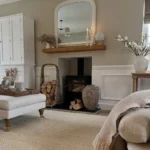 A rustic living room in neutral-tone with a wood-burning stove, stacked firewood, a large mirror above the mantel, and cozy decor including a rug, vases, and candles.