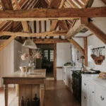 A rustic kitchen with exposed wooden beams, a wooden island with vases, white cabinets, a black stove, and hanging utensils on a white tile wall—perfect for inspiring your rustic kitchen ideas.