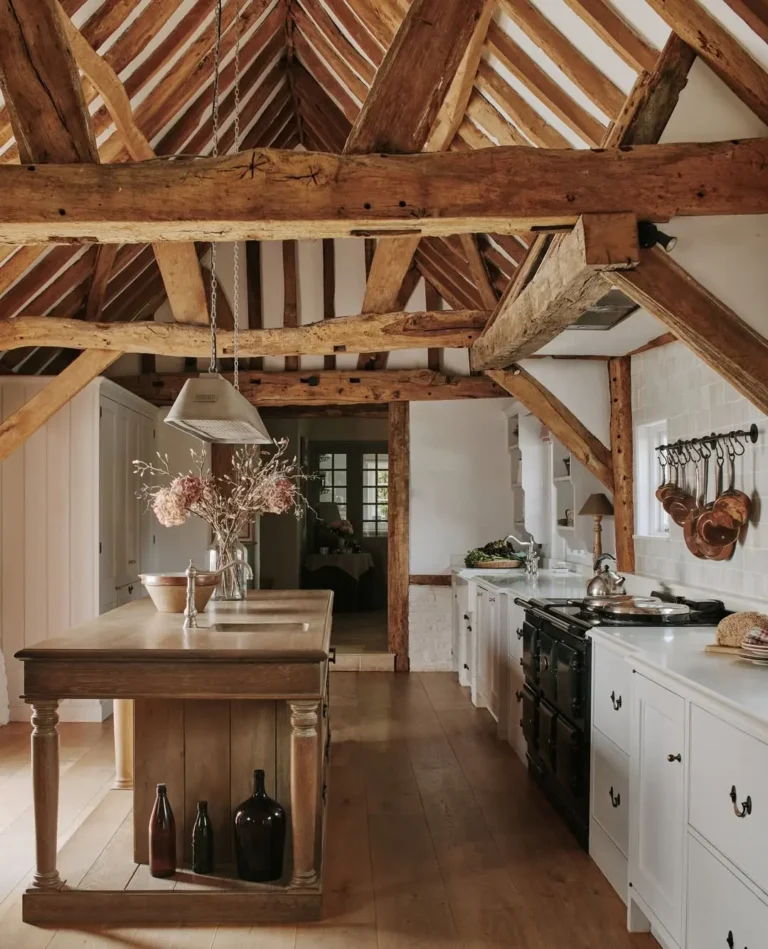 A rustic kitchen with exposed wooden beams, a wooden island with vases, white cabinets, a black stove, and hanging utensils on a white tile wall—perfect for inspiring your rustic kitchen ideas.