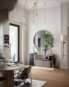 Entryway with stylish modern decor, featuring a round mirror, bench, plants, decorative objects, and pendant lights. Light wood floor, neutral walls, and a dining area with table and chairs complete the look.