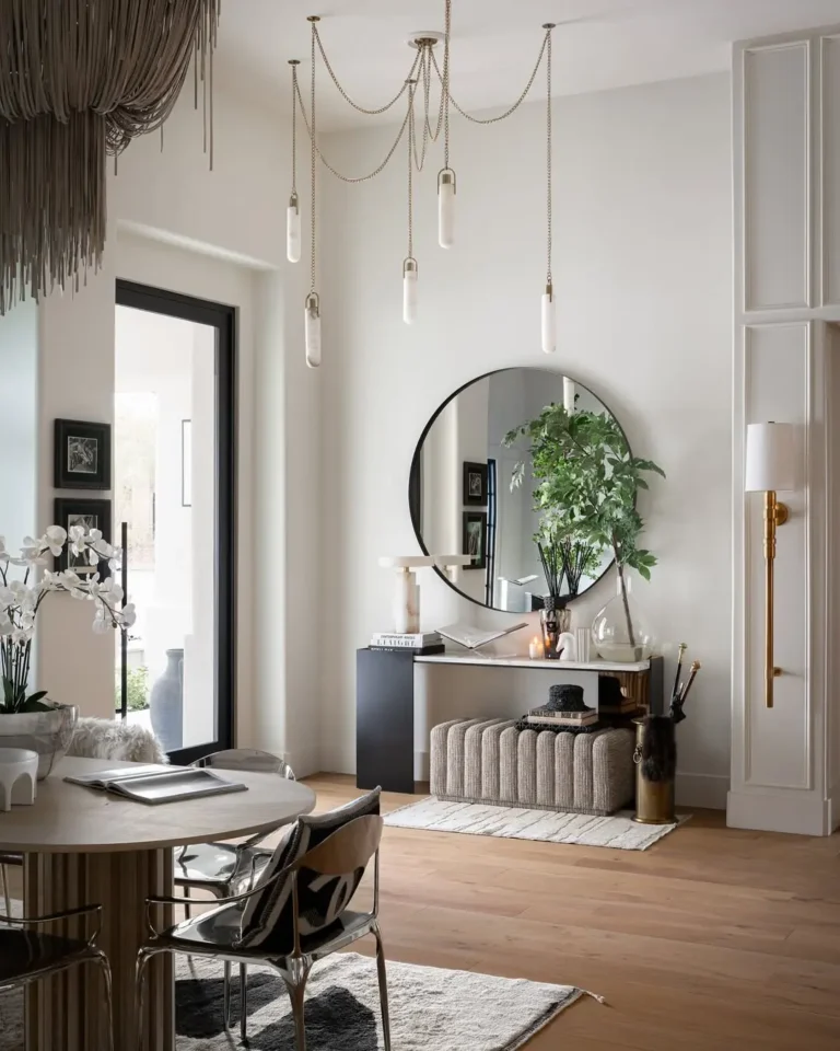Entryway with stylish modern decor, featuring a round mirror, bench, plants, decorative objects, and pendant lights. Light wood floor, neutral walls, and a dining area with table and chairs complete the look.