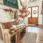 A farmhouse-style entryway with a wooden bench, autumn decorations, throw blankets, a chalkboard sign, and double wooden doors—your perfect inspiration from our farmhouse fall decor guide.