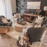A cozy farmhouse fall living room decor with gray armchairs, a round tufted ottoman, woven rug, and wood accents, featuring decorative pillows, plants, and a sideboard against a dark paneled wall.