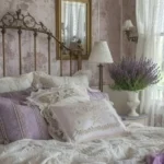 A vintage-style bedroom with a brass headboard, floral wallpaper, lace-trimmed pillows and bedding in lavender and white, a vase of lavender, and a mirror above a nightstand evokes classic french country color palettes.