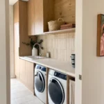 Modern laundry room with wood cabinets, a countertop, a sink, two front-loading machines, beige tiled backsplash, and shelves holding baskets and towels—thoughtfully arranged to avoid common laundry room design mistakes.
