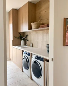 Modern laundry room with wood cabinets, a countertop, a sink, two front-loading machines, beige tiled backsplash, and shelves holding baskets and towels—thoughtfully arranged to avoid common laundry room design mistakes.