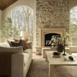 A neutral-toned living room with a stone fireplace, large arched windows, a beige sofa, a light coffee table, and decorative plants and candles highlights the beauty of natural materials in home decor.