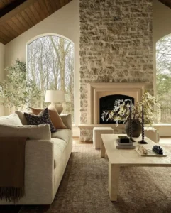 A neutral-toned living room with a stone fireplace, large arched windows, a beige sofa, a light coffee table, and decorative plants and candles highlights the beauty of natural materials in home decor.