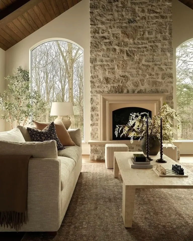 A neutral-toned living room with a stone fireplace, large arched windows, a beige sofa, a light coffee table, and decorative plants and candles highlights the beauty of natural materials in home decor.