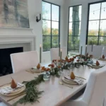 A dining table set for six with white chairs, gold cutlery, striped napkins, pears on plates, candles, and greenery centerpieces in a bright room with large windows—perfect for minimalist Thanksgiving decor ideas.