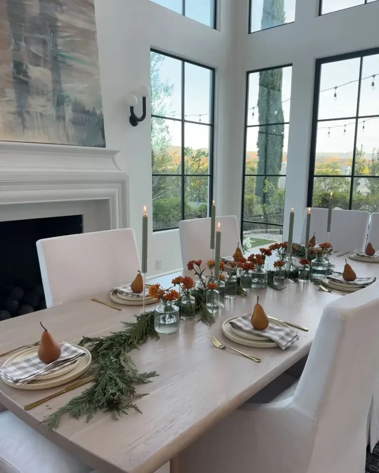 A dining table set for six with white chairs, gold cutlery, striped napkins, pears on plates, candles, and greenery centerpieces in a bright room with large windows—perfect for minimalist Thanksgiving decor ideas.