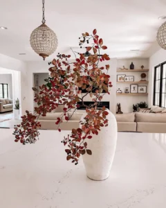 A white vase with branches of red and brown leaves sits on a marble countertop in a modern, well-lit living room—perfect inspiration from a modern fall decor guide.