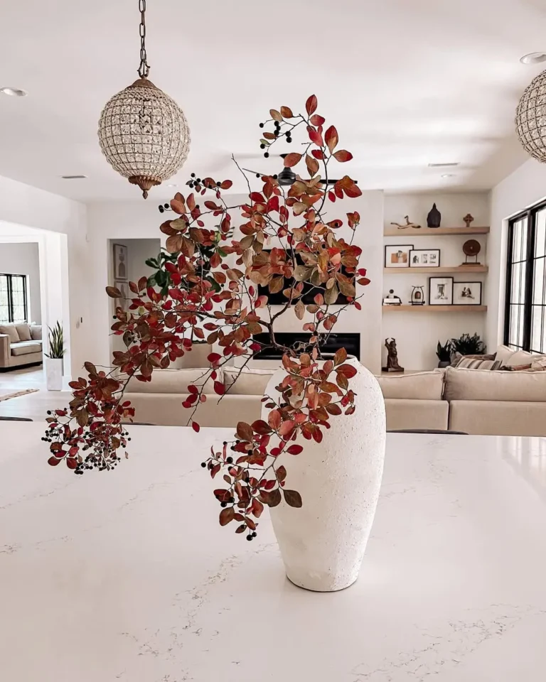 A white vase with branches of red and brown leaves sits on a marble countertop in a modern, well-lit living room—perfect inspiration from a modern fall decor guide.