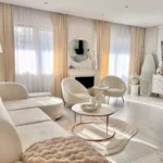 A modern living room inspired by a modern home decor guide features white and beige furniture, sheer curtains, a round coffee table, a circular mirror, and chic accents in a bright, sunlit space.