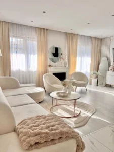 A modern living room inspired by a modern home decor guide features white and beige furniture, sheer curtains, a round coffee table, a circular mirror, and chic accents in a bright, sunlit space.
