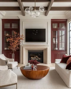 A modern living room with a fireplace, wall-mounted TV, red built-in shelves, neutral sofas, a round wooden coffee table, and organic modern fall decor like decorative vases and plants.