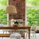 Rustic dining room decor with a wood table, pale green spindle chairs, a brick fireplace, and a woven pendant light—set against large windows and a forest view.