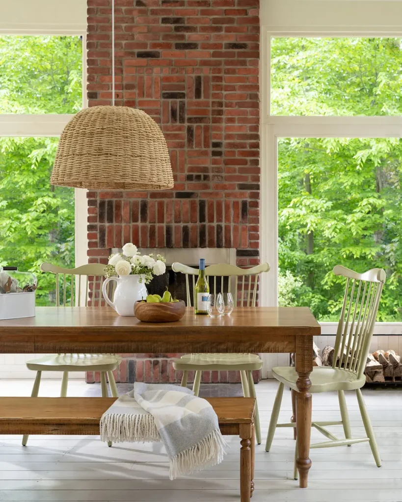 Rustic dining room decor with a wood table, pale green spindle chairs, a brick fireplace, and a woven pendant light—set against large windows and a forest view.
