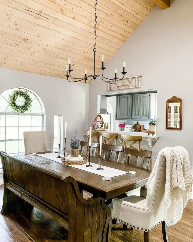 A dining room with a wooden table, bench seating, upholstered chairs, rustic dining room lighting chandelier, kitchen pass-through, barstools, and a large arched window with a wreath.