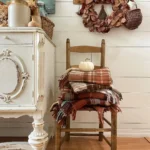 A wooden chair holds folded plaid blankets and a white pumpkin, next to a white vintage cabinet, with rustic fall decor like a brown wreath and basket hanging on a white shiplap wall.