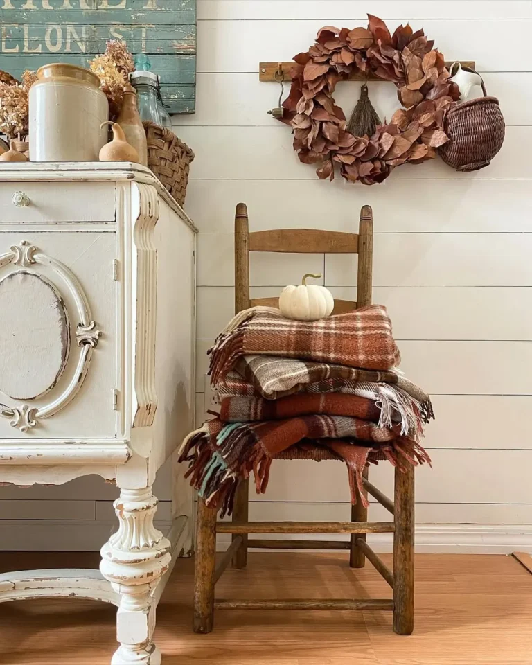 A wooden chair holds folded plaid blankets and a white pumpkin, next to a white vintage cabinet, with rustic fall decor like a brown wreath and basket hanging on a white shiplap wall.