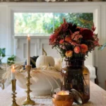 A rustic fall dining table centerpiece with a wooden table, two brass candlesticks, a lit candle in a copper holder, a vase of autumn flowers, a white bowl with a white pumpkin, and wheat stalks.