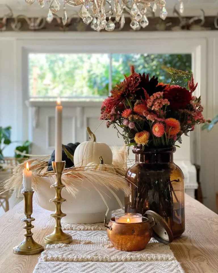 A rustic fall dining table centerpiece with a wooden table, two brass candlesticks, a lit candle in a copper holder, a vase of autumn flowers, a white bowl with a white pumpkin, and wheat stalks.