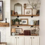 Built-in shelves with books, framed artwork, and rustic seasonal decor ideas like vases with flowers and decorative objects arranged above white cabinets in a living room.