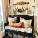 A black wooden bench with beige cushions and a "be thankful" pillow sits against a wall adorned with hanging gold bells, wicker baskets underneath—a perfect touch of Thanksgiving entryway decor.