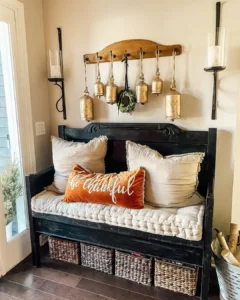 A black wooden bench with beige cushions and a "be thankful" pillow sits against a wall adorned with hanging gold bells, wicker baskets underneath—a perfect touch of Thanksgiving entryway decor.