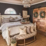 A cozy farmhouse fall bedroom with a large bed, neutral bedding, a tufted bench, wooden sliding doors adorned with wreaths, a jute rug, and a sparkling crystal chandelier.