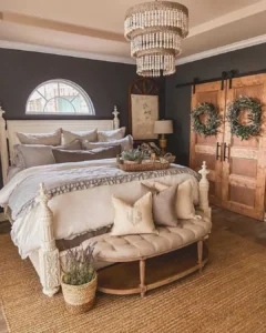 A cozy farmhouse fall bedroom with a large bed, neutral bedding, a tufted bench, wooden sliding doors adorned with wreaths, a jute rug, and a sparkling crystal chandelier.