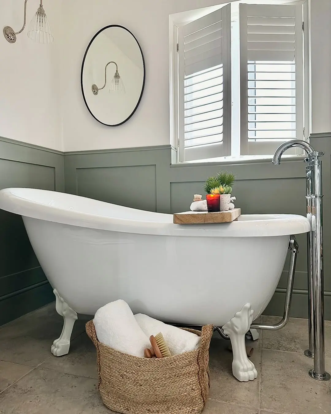 Clawfoot roll top bathtub with chrome faucet styled for a farmhouse bathroom look, featuring green board and batten walls, round mirror, and woven basket with towels.