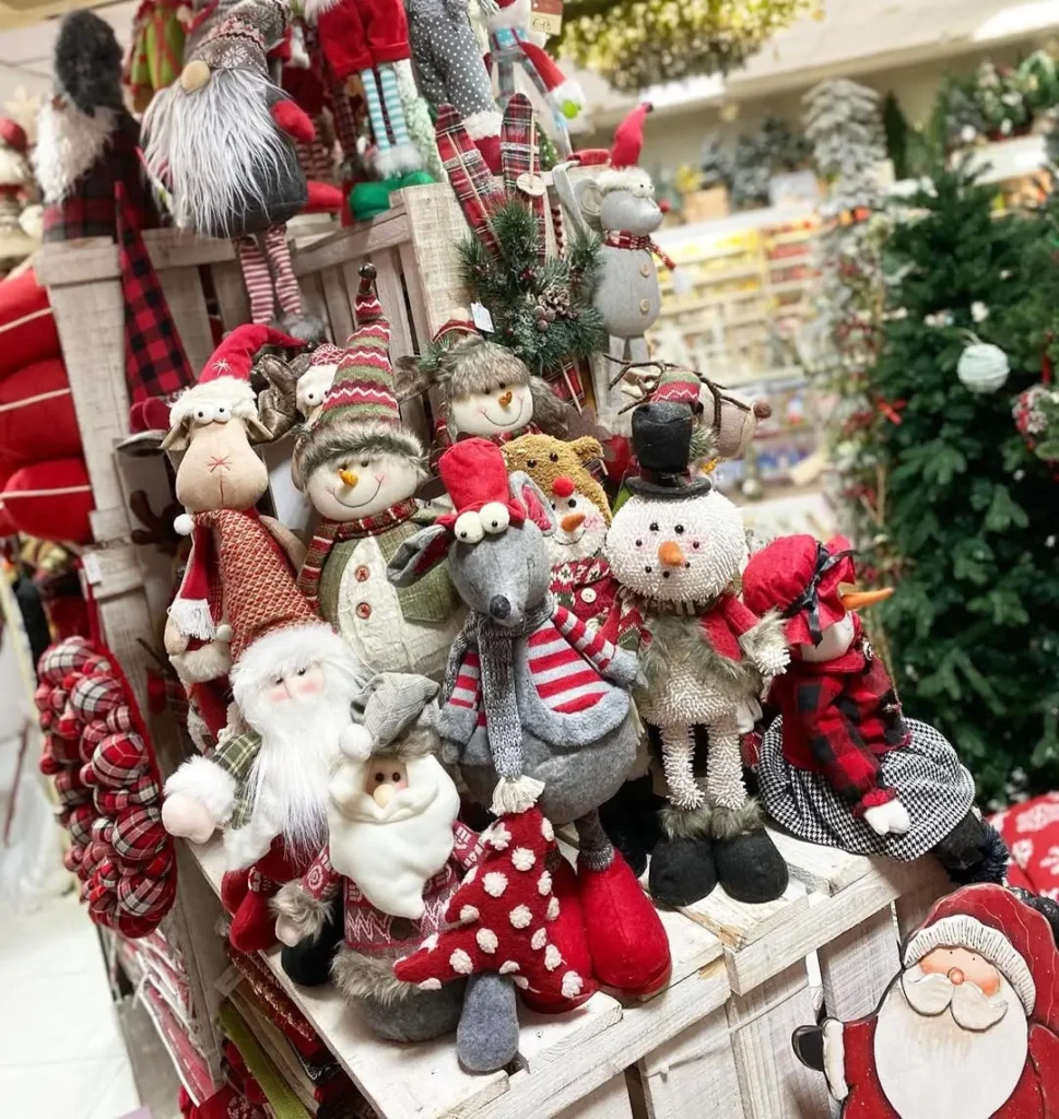A display of Christmas character plush toys, including snowmen, Santa Claus, reindeer, and gnomes, arranged on wooden crates in a store.