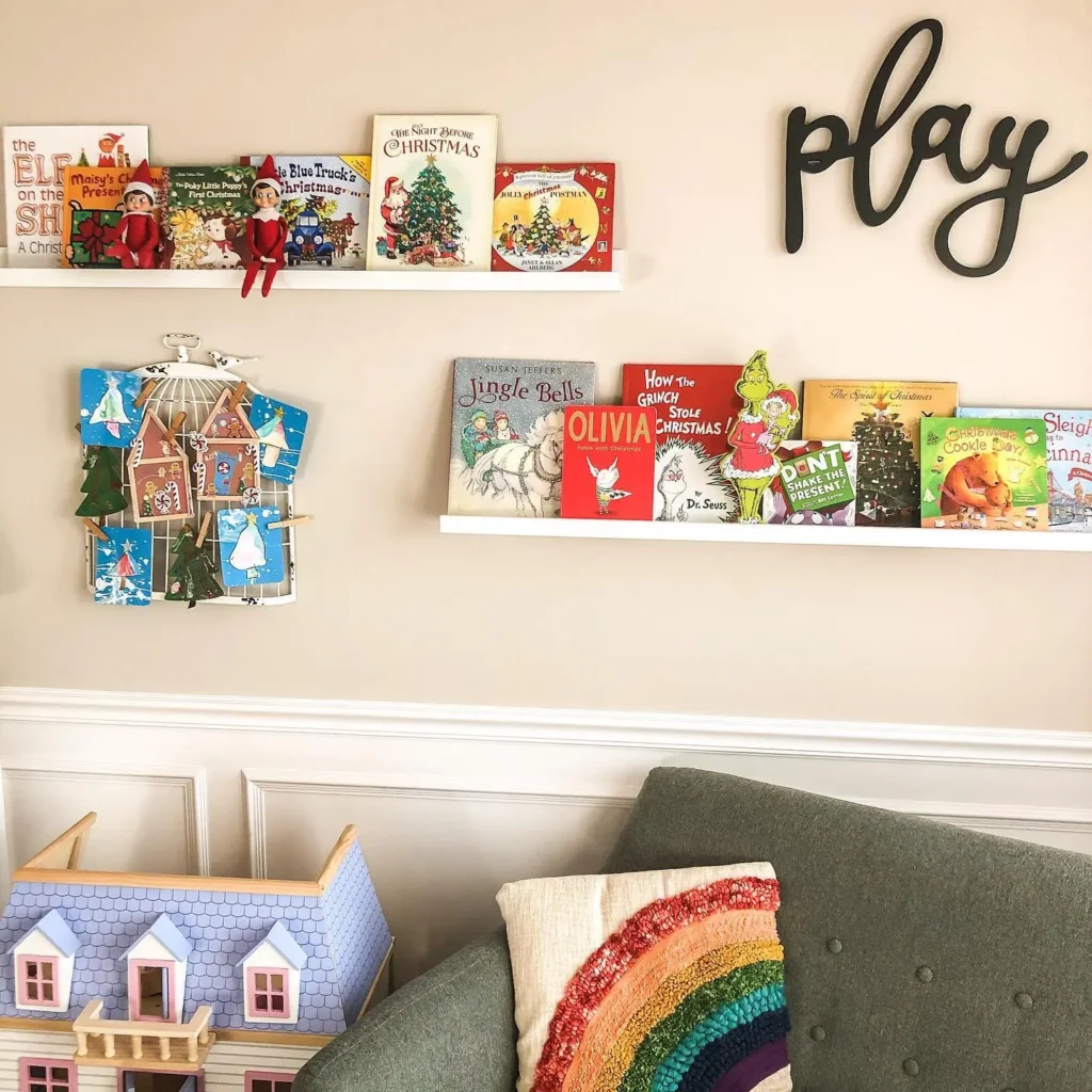 A playroom features a Christmas kids storybook display on shelves, two elf dolls, a book-themed advent calendar, a green couch with a rainbow pillow, and a dollhouse. The word "play" decorates the wall.