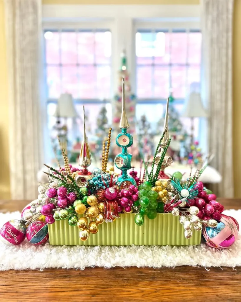 A festive Christmas table centerpiece for kids features colorful ornaments, beads, and baubles in a green rectangular container, sitting on a table in front of a bright window.
