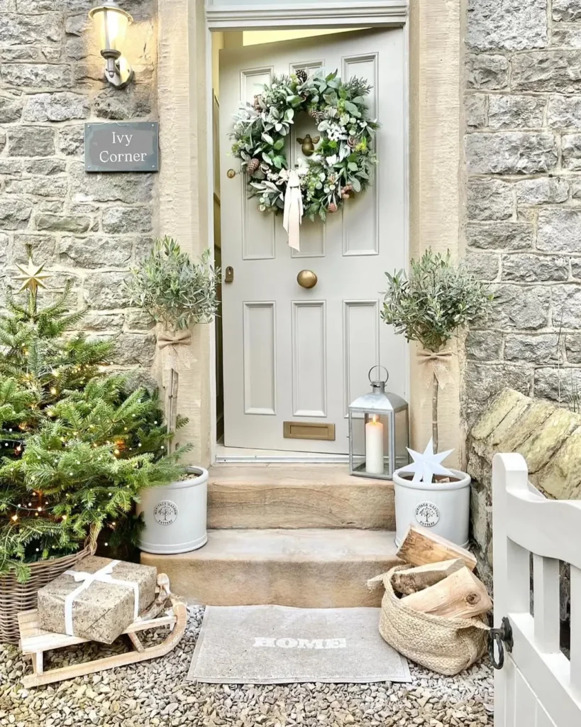 A front door with festive Christmas entryway decor: a green wreath, potted plants, a Christmas tree on a sled, wrapped gifts, a lantern, and a "HOME" doormat against a stone wall.
